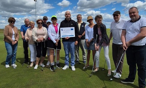 Grupa kilkunastu uczestników stoi na polu golfowym podczas zajęć sportowych realizowanych w ramach projektu. Część osób trzyma kije golfowe, a jedna osoba prezentuje tablicę informacyjną dotyczącą dofinansowania przedsięwzięcia ze środków Funduszy Europej
