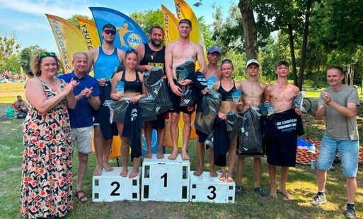 Uczestnicy turnieju siatkówki plażowej stoją na podium podczas ceremonii wręczenia nagród. Zawodnicy trzymają puchary, upominki i koszulki, a obok nich stoją organizatorzy. W tle widoczne są flagi wydarzenia oraz teren rekreacyjny wśród drzew.