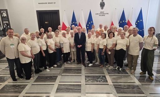 Grupowe zdjęcie osób stojących w reprezentacyjnej sali Senatu Rzeczypospolitej Polskiej. W centrum znajduje się mężczyzna w granatowym garniturze, otoczony przez grupę głównie starszych kobiet i mężczyzn ubranych w jasne koszulki. W tle widoczne są flagi 