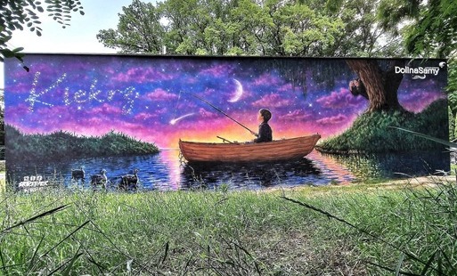 Mural na ścianie Świetlicy Wiejskiej w Kiekrzu przedstawiający jezioro, łódkę z wędkującym dzieckiem, ptaki oraz rozgwieżdżone niebo z księżycem. Dzieło otoczone jest zielenią i drzewami.