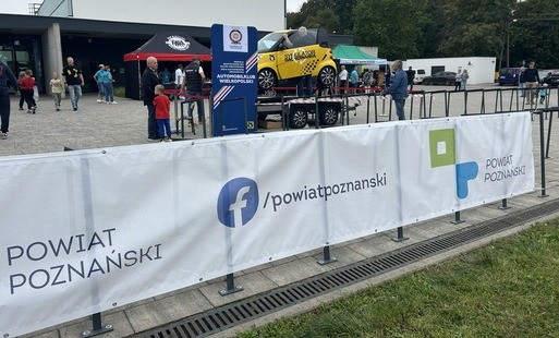 Zdjęcie przedstawia plenerowe wydarzenie motoryzacyjne przed budynkiem. Na pierwszym planie znajduje się biała barierka z logo i napisem „Powiat Poznański” oraz adresem „/powiatpoznanski” na Facebooku. W centrum stoi żółty samochód wystawiony na przyczepie, z napisem „Szkoła Dachowania”, obok dużej tablicy „Automobilklub Wielkopolski”. Wokół znajdują się namioty wystawców, zwiedzający oraz zaparkowane samochody.